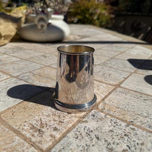 May include: A small, polished silver cup with a rounded base and a smooth, cylindrical body. The cup reflects the surrounding environment, creating a mirrored effect. The cup is sitting on a stone tile surface.