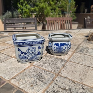 Könnte beinhalten: Zwei kleine, quadratische, weiße Keramiktöpfe mit blauen Blumenmustern. Der größere Topf hat ein Blumen- und Blättermuster an der Seite, während der kleinere Topf ein aufwendigeres Design hat. Beide Töpfe haben einen blauen Rand oben.