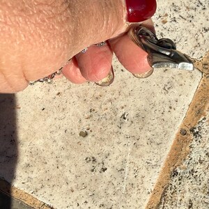 May include: A silver-toned pendant with a stylized figure, possibly a dancer or a person in motion. The pendant is attached to a delicate chain. The background is a textured, light-colored surface.