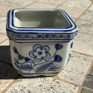 Könnte beinhalten: Kleiner, quadratischer, weißer Keramiktopf mit blauen Blumenmustern. Der Topf hat einen blauen Rand und eine dekorative Bordüre. Das Blumenmuster umfasst Blumen, Blätter und Stiele. Der Topf ist wahrscheinlich für kleine Pflanzen oder Sukkulenten.