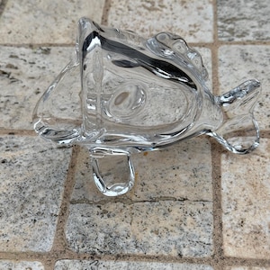 Puede incluir: Un objeto decorativo transparente en forma de pez de cristal. El pez tiene aletas detalladas y un lazo en la parte inferior. El objeto descansa sobre una superficie de baldosas de piedra. El cristal es transparente.