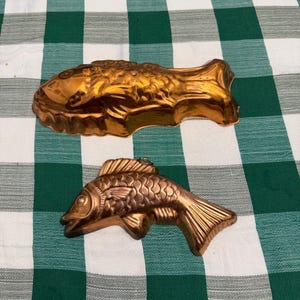 May include: Two copper-colored fish molds. One is a larger, detailed fish shape, and the other is a smaller, more stylized fish. Both are made of metal and are likely used for baking or decorative purposes. The molds are on a green and white checkered tablecloth.