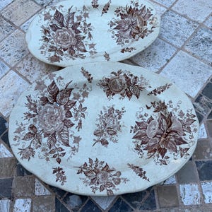 May include: Two oval-shaped antique serving platters. The off-white platters feature a brown floral pattern with roses and other flowers. The floral design is spread across the surface of the platters, with a detailed arrangement of leaves and blossoms.