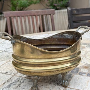 Könnte beinhalten: Ein antiker Pflanztopf aus Messing mit geschwungenem Rand und zwei verzierten Griffen. Der Pflanztopf hat ein geripptes Design und steht auf drei Klauenfüßen. Das Messing hat eine warme, gealterte Patina, ein dekorativer Gegenstand.
