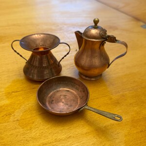 Puede incluir: Un juego de tres utensilios de cocina de cobre en miniatura: una jarrita pequeña, una taza en forma de embudo y una sartén.