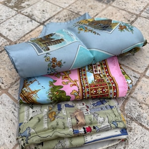 May include: A stack of silk scarves with travel-themed designs. The top scarf is light blue with a print of a building and floral accents. Below it is a pink scarf with a red and gold border. The bottom scarf is green with a map-like design.