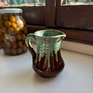 Può includere: Un piccolo brocca in ceramica marrone e verde con un manico. La brocca ha una finitura smaltata e un labbro leggermente svasato.