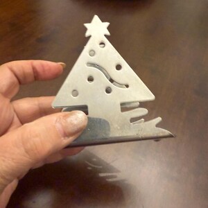 Op de afbeelding: Een zilverkleurige metalen kerstboom-onderzetter met een ster erbovenop. De onderzetter heeft decoratieve uitgesneden gaten en een golvend lijnenontwerp. De onderzetter wordt door een hand vastgehouden, met een stapel blauw-witte borden op de achtergrond.