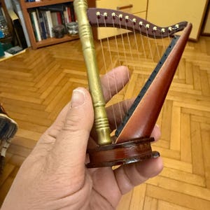 May include: Miniature harp with a wooden frame and gold-colored accents. The strings are visible, and the instrument is held in a hand. The harp is a musical instrument.