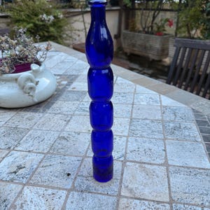 Könnte beinhalten: Eine hohe, kobaltblaue Glasflasche mit einem einzigartigen, gestapelten Design. Die Flasche besteht aus einer Reihe von abgerundeten Abschnitten, die eine interessante Form ergeben. Die Flasche steht auf einer gefliesten Oberfläche und zeigt ihre lebendige Farbe.