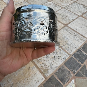 Może przedstawiać: Srebrny metalowy świecznik z wytłoczonym wzorem winorośli. Świecznik ma wycięte wzory i okrągłą podstawę.