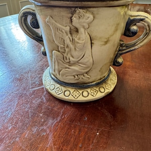 Peut inclure: Un pot en céramique avec un motif en relief d'une femme jouant de la lyre. Le pot a deux anses et une base avec un motif géométrique. Le pot est beige avec des accents bleus.
