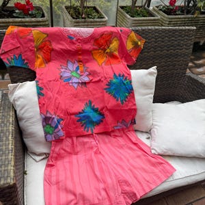 May include: A two-piece pink pajama set with a floral print. The short-sleeved top has a button-up neckline and is adorned with large, colorful flowers in shades of orange, yellow, blue, and purple. The matching shorts are a solid pink with vertical stripes.