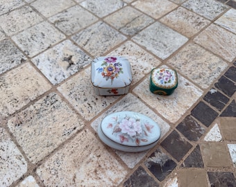 May include: Three decorative porcelain trinket boxes with floral designs. The boxes are white, green, and oval-shaped, with colorful flower patterns on the lids. The boxes are placed on a stone tile surface.