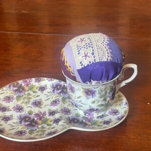 Op de afbeelding: Een paars en wit stoffen kussen zit in een bloemetjes theekopje en schoteltje. Het schoteltje heeft een bloemetjespatroon met paarse viooltjes.