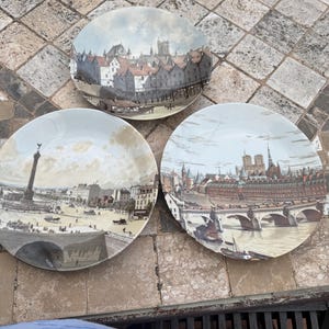 May include: Three decorative plates featuring vintage city scene illustrations. The plates depict European cityscapes, including buildings, bridges, and figures. The plates are round and have a white background with a detailed illustration.