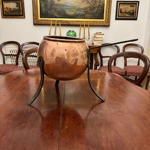 Può includere: Una pentola di rame con finitura martellata si trova su un treppiede. La pentola è su un tavolo di legno in una stanza con quadri alle pareti.