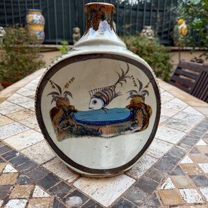 Op de afbeelding: Een keramische vaas met een rond lichaam en een smalle hals. De vaas is versierd met een bruine glazuur en een handgeschilderd ontwerp van een vis die in een blauwe vijver zwemt. De vis is bruin, wit en blauw.