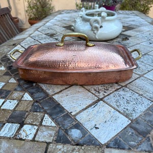 Puede incluir: Una asadera de cobre martillado con tapa y asas de latón. La bandeja rectangular se exhibe sobre una mesa de piedra. La tapa tiene un asa de latón. La bandeja está diseñada para cocinar y hornear.
