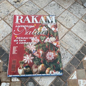 May include: A magazine titled "RAKAM" with the word "Natale" prominently displayed. The cover features a Christmas tree adorned with ornaments. Text includes "REGALI IDEA da fare e comprare" and "CASA DECORATA RICAMO CLASSICO NUOVI DISEGNI PUNTO CROCE MAGLIA PREZIOSA."