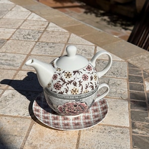 Può includere: Un servizio da tè in ceramica bianca con motivi floreali, composto da una teiera, una tazza e un piattino. La teiera e la tazza sono impilate. Il piattino e il servizio da tè presentano un motivo floreale rosa e marrone. Il testo "MATHILDE M. - FRANCE" è visibile.