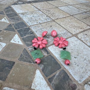 May include: A pair of pink flower-shaped earrings with green square and pink round accents. The earrings are displayed on a multi-colored stone tile surface. The earrings have a post-back closure.