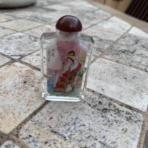 May include: A small, clear glass perfume bottle with a brown stopper. The bottle features a painted scene of figures in traditional clothing. The artwork includes shades of pink, blue, and gold. The bottle is rectangular with a rounded top.