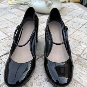 May include: A pair of black patent leather Mary Jane pumps with a glossy finish, featuring a rounded toe, a thin adjustable strap across the instep, and a block heel. The interior footbed is light beige and displays the Aldo brand name clearly.