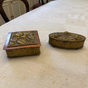 Puede incluir: Dos cajas antiguas de bronce con diseños florales en relieve. Una es rectangular con un marco color cobre, y la otra es ovalada. Ambas cajas están sobre un mantel crema, mostrando detalles intrincados y una estética vintage.