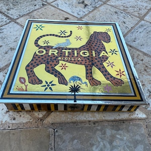 Peut inclure: Boîte rectangulaire avec un design or et noir. La boîte présente une grande illustration de léopard avec le texte "ORTIGIA SICILIA". Le fond est jaune avec des accents floraux et étoilés.