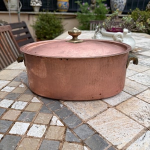 May include: A copper pot with a lid and brass handles. The pot is round with a hammered texture and a brass knob on the lid. The pot is sitting on a stone table outdoors.
