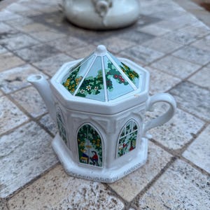 Può includere: Teiera in ceramica bianca con un design a serra. Il coperchio presenta un motivo floreale su sfondo azzurro. I lati della teiera raffigurano finestre ad arco con figure e vegetazione. La parola "conservatory" è stampata sulla base.