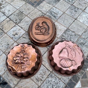 Puede incluir: Tres moldes antiguos de cobre con diseños detallados. Uno presenta una ardilla, otro una flor y el tercero una pera y una piña. Cada molde tiene un lazo para colgar y está sobre una superficie de baldosas estampadas.