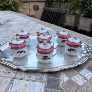 May include: A silver serving tray holds six small, white porcelain cups with lids. Each cup features a pink band, gold trim, and a bird illustration. The lids are white with a small floral knob. The cups are arranged on a silver tray.