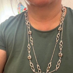 May include: Silver-toned necklace with a double-strand design. The necklace features decorative knot-shaped accents and intricate chain links. The jewelry is worn against a green shirt.