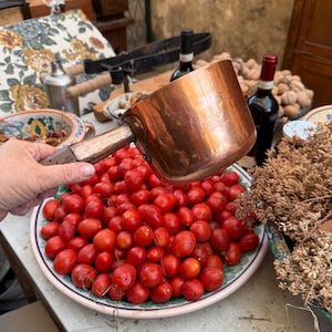 May include: A copper pot with a wooden handle is being used to scoop up a bowl of red tomatoes. The tomatoes are in a white bowl with a blue and green floral pattern.