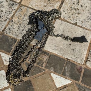 May include: A silver-toned necklace with multiple charms, including a butterfly and a bat. The chain is made of small, interconnected links. The necklace is lying on a stone surface.