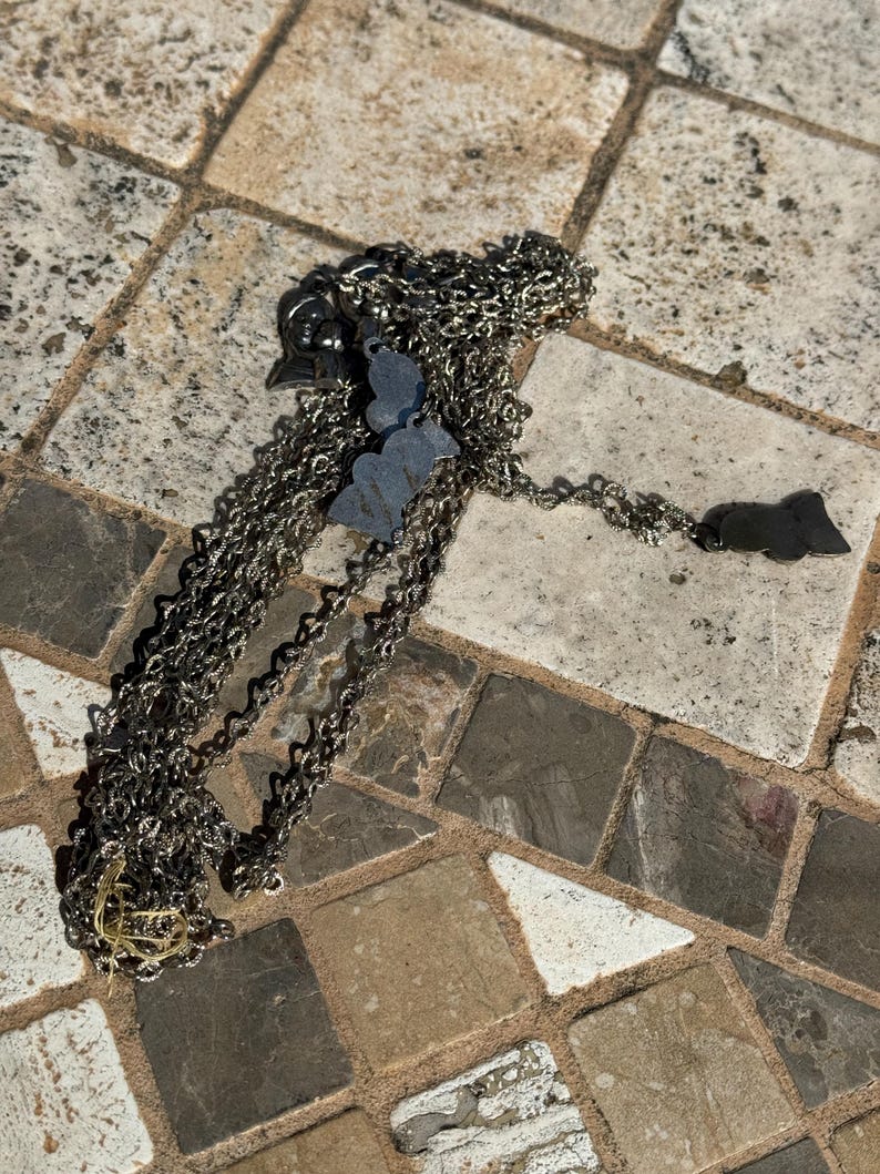 May include: A silver-toned necklace with multiple charms, including a butterfly and a bat. The chain is made of small, interconnected links. The necklace is lying on a stone surface.