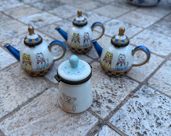 Puede incluir: Colección de miniaturas de cerámica: teteras y un pequeño tarro con tapa. Las teteras son blancas con detalles azules, ribetes dorados e ilustraciones de gatos. El tarro es blanco con una ilustración de oso de peluche y tapa celeste.