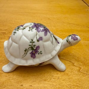 Può includere: Una statuetta di tartaruga in ceramica bianca con un design floreale viola e verde. La tartaruga ha un guscio dettagliato e quattro zampe.