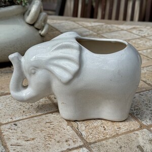 May include: White ceramic elephant planter with a small opening at the top for planting. The planter is sitting on a tiled surface.