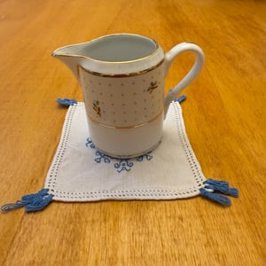 May include: A white ceramic creamer with a gold trim and a blue and white floral pattern. The creamer is sitting on a white crocheted coaster with blue tassels.
