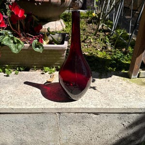 May include: A tall, teardrop-shaped, ruby-red glass vase. The vase has a long, slender neck and a rounded base. The vase is sitting on a concrete surface, with a garden in the background.