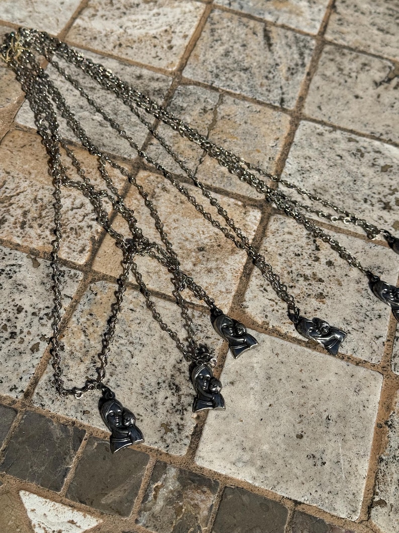 May include: Several silver-toned necklaces with pendant charms featuring a mother and child design. The necklaces are laid out on a stone tile surface, showcasing the intricate details of the pendants and chains.