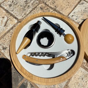 Pode incluir: Um conjunto de acessórios para vinho numa caixa redonda de bambu. O conjunto inclui um saca-rolhas, um bico e dois tampões. O saca-rolhas e o bico têm cabos de madeira. Os tampões são prateados e de madeira. O interior da caixa é branco.