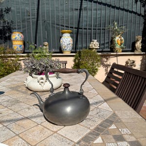 May include: A pewter teapot with a wooden handle sits on a tiled table in an outdoor setting. The teapot has a round body and a spout on the side. The table is made of small, square tiles in shades of beige and gray.