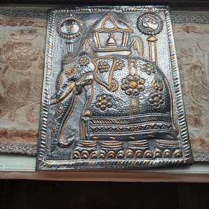 May include: A rectangular metal wall plaque featuring a silver and gold embossed elephant design. The elephant is adorned with floral patterns and carries a canopy on its back, flanked by circular sun and moon motifs within a decorative border.