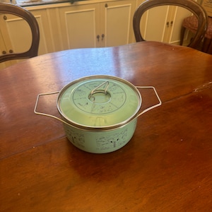 May include: A light green enamelware pot with a lid and a silver handle. The pot has a silver rim and a silver handle. The lid has a silver handle. The pot has a black illustration around the sides. The pot is on a wooden table.