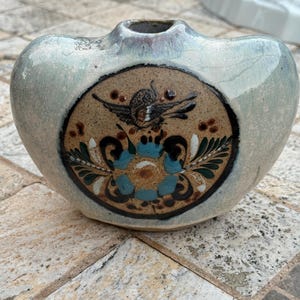 May include: A ceramic vase with a heart-shaped design and a light blue and gray glaze. The front features a circular design with a bird and floral pattern in brown, blue, and black. The vase has a small opening at the top.