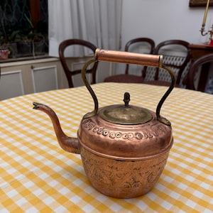 Può includere: Una teiera di rame con un coperchio e un manico in ottone. La teiera ha un design elaborato e si trova su una tovaglia a quadri gialla e bianca.